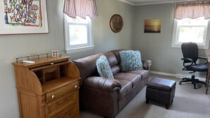 Una sala de estar con un sofá de cuero marrón, una mesita de madera, una silla de oficina negra y un ventanal con cortinas.