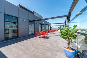 A rooftop terrace with red chairs and tables, a potted plant, and a glass-enclosed building with a view of the city.