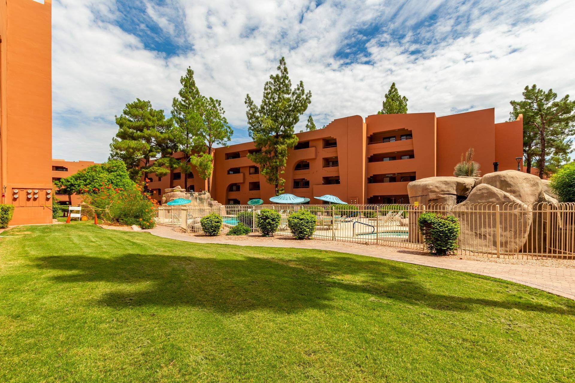 A hotel with a modern orange facade, multiple balconies, and a well-maintained lawn with trees and shrubs.
