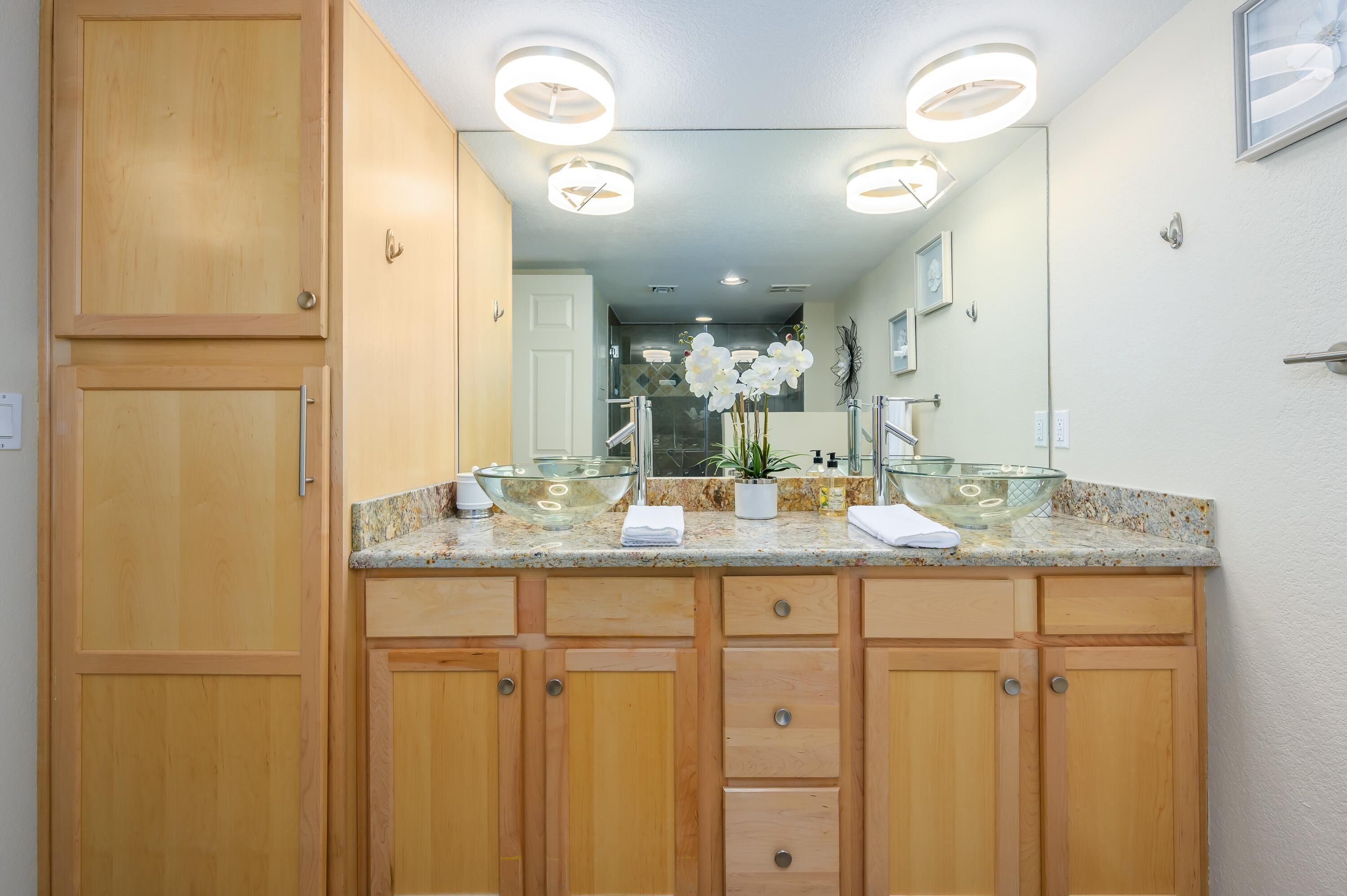 A bathroom with wooden cabinets, a glass bowl sink, and a large mirror with overhead lighting.