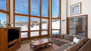 Ein Wohnzimmer mit großen Fenstern, einem Fernseher und einem Sofa.