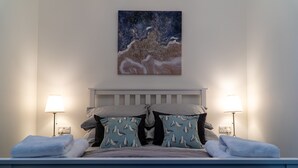 Un dormitorio con cama, almohadas, una mesita de noche, una lámpara y un cuadro en la pared.