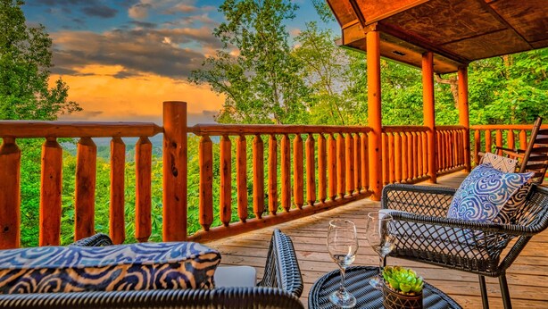 A wooden deck with wicker furniture and a glass table, overlooking a lush green landscape. - Dollywood – HotTub,Theater,Pool,BBQ,Arcade & Views (Sevierville)