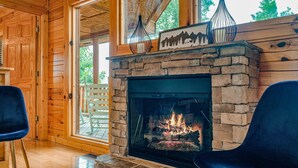 A cozy room with a stone fireplace, wooden walls, and a large window. - Dollywood – HotTub,Theater,Pool,BBQ,Arcade & Views (Sevierville)