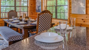 A wooden cabin interior with a dining table set for four, chairs with patterned upholstery, and large windows with blinds. - Dollywood – HotTub,Theater,Pool,BBQ,Arcade & Views (Sevierville)