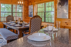 A wooden cabin interior with a dining table set for four, chairs with patterned upholstery, and large windows with blinds.