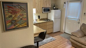 A compact kitchen with wooden cabinets, a microwave, a refrigerator, and a dining area with a chair.