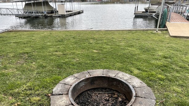 A circular stone fire pit in a grassy area with a body of water and docks in the background.