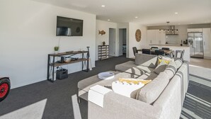 En moderne stue med sofa, sofabord og fjernsyn ophængt på væggen. Der er et køkkenområde med køleskab og spisebord.