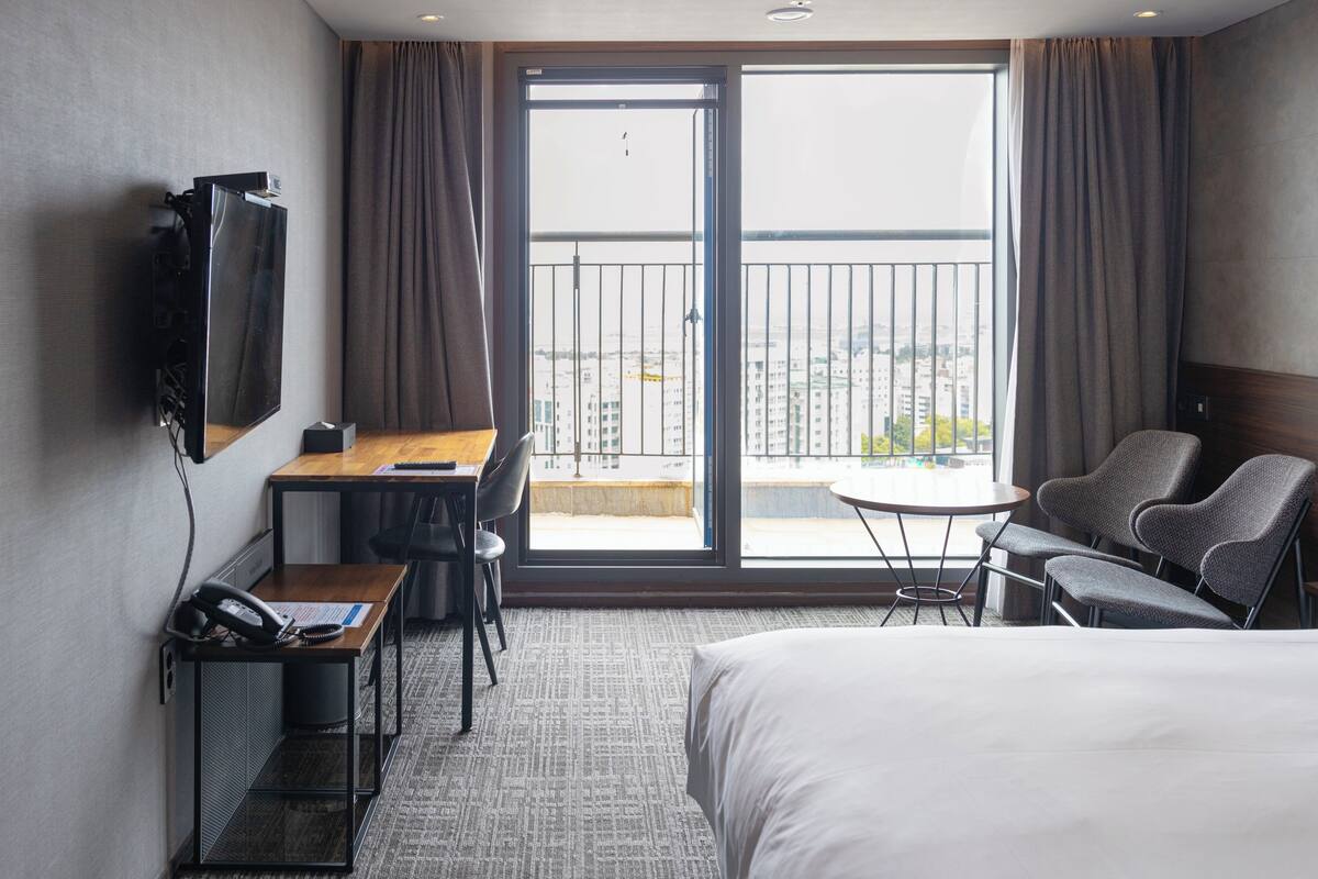 A hotel room with a bed, a desk with a chair, a small table with two chairs, a TV mounted on the wall, and a balcony with a view of the city.
