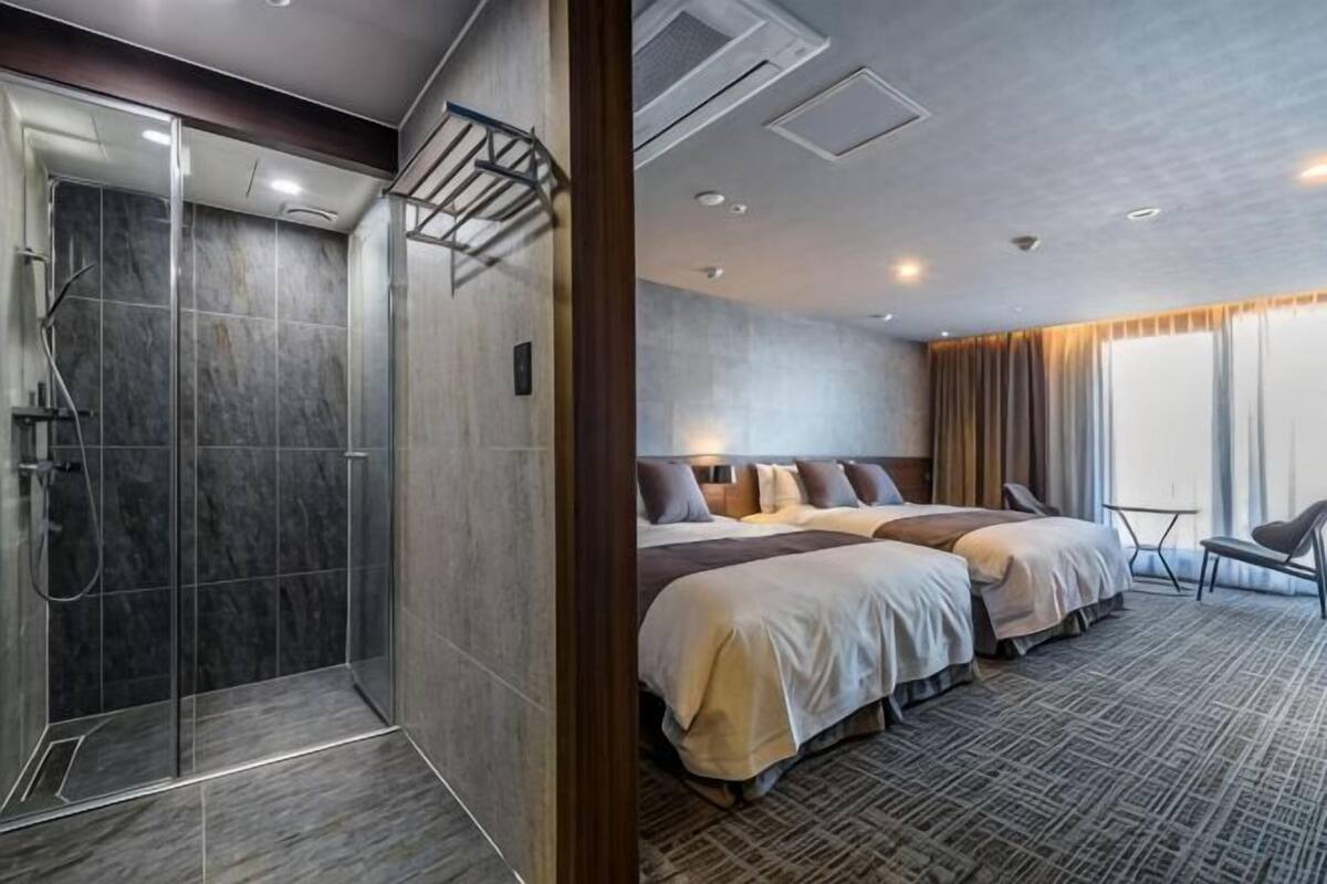 A hotel room with a shower, two beds, a sitting area, and a large window.
