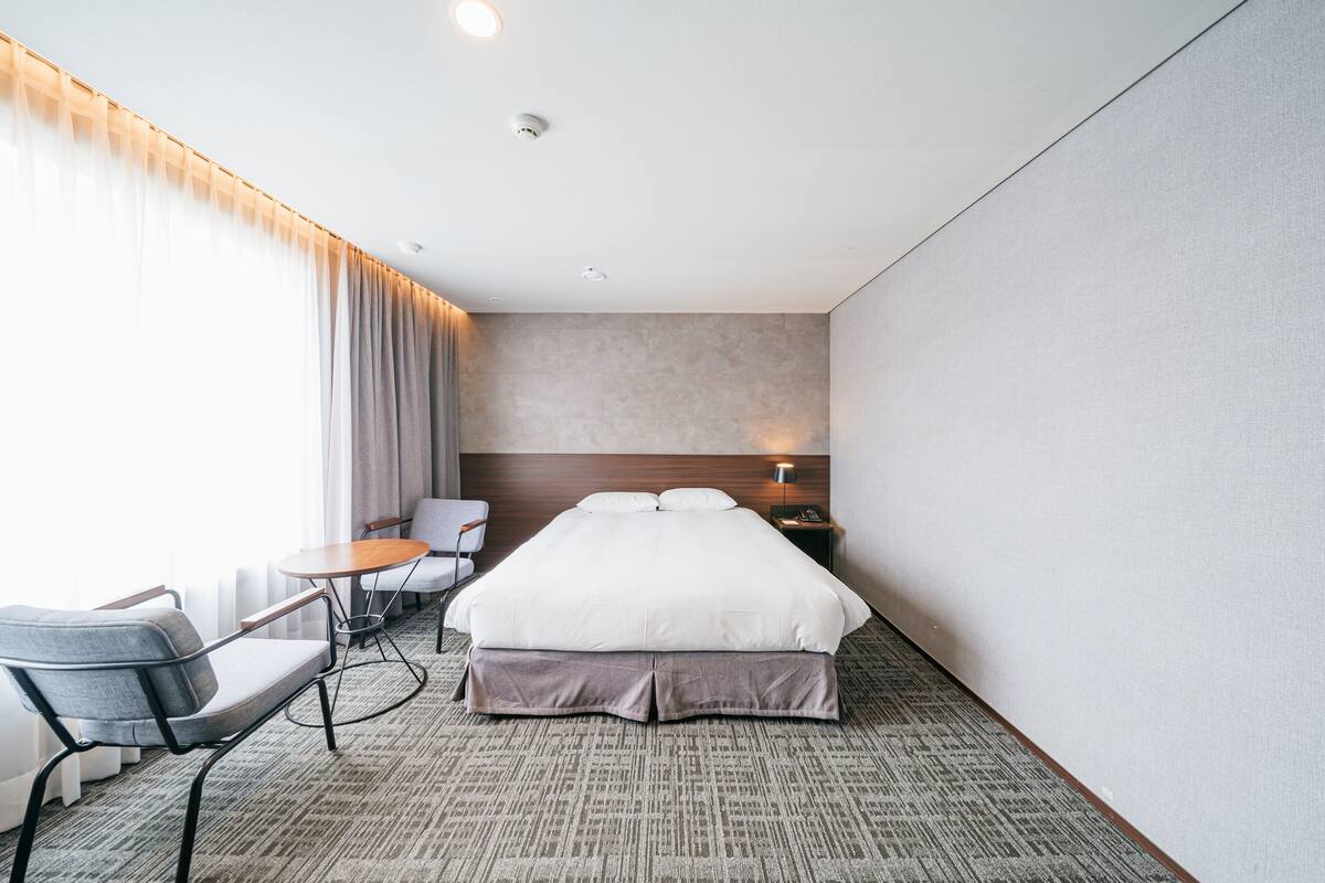 A hotel room with a bed, a desk, a chair, a lamp, and a window with curtains.