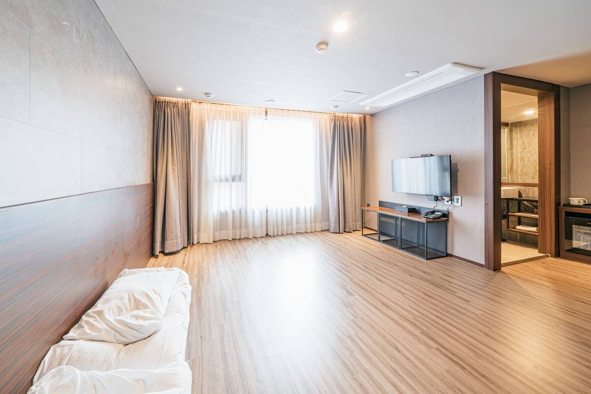 A modern hotel room with a wooden floor, a bed, a desk with a chair, a television, and a bathroom visible through an open door.