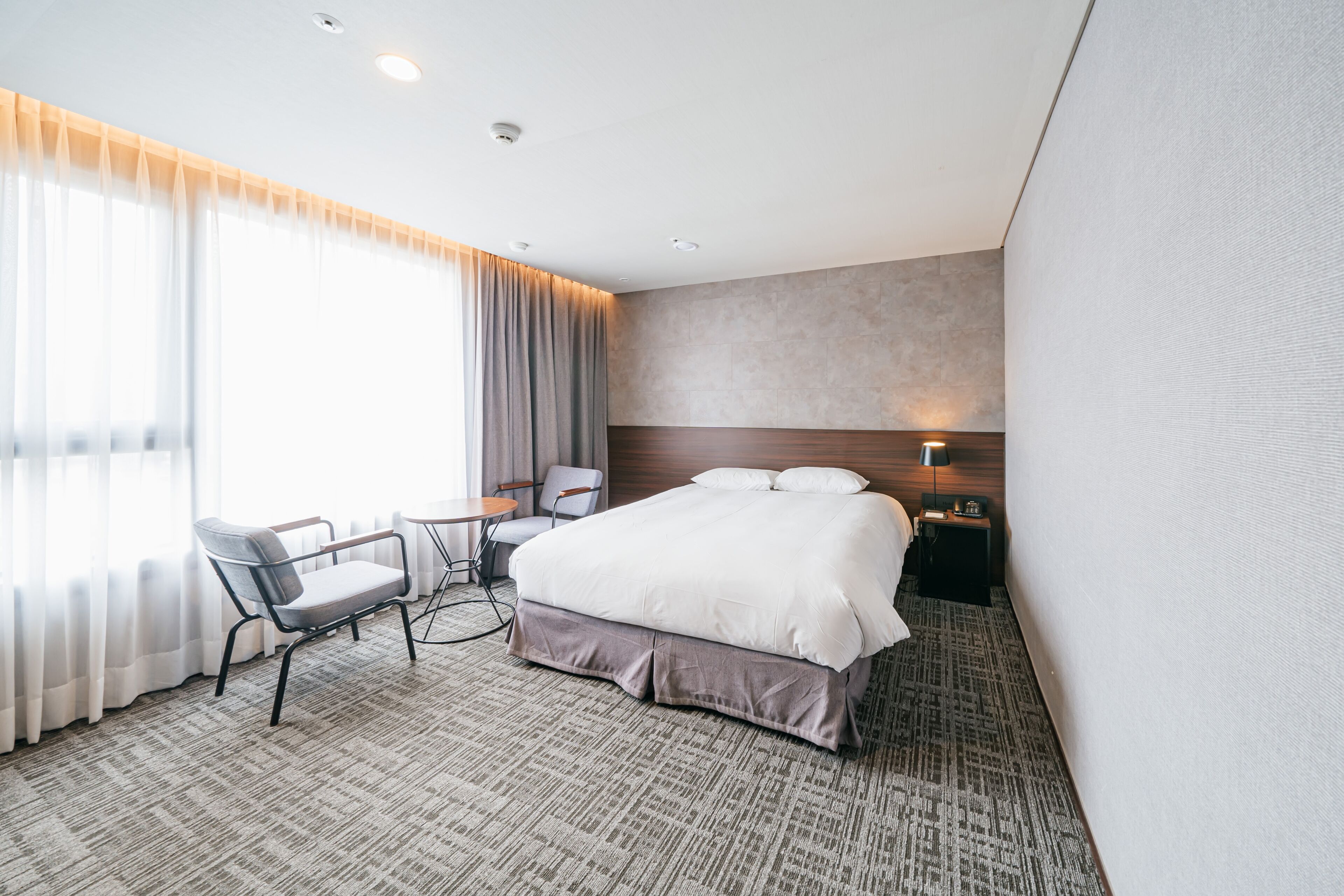 Habitación de hotel con una cama grande, un escritorio con silla, una mesita pequeña y una mesita de noche con lámpara.