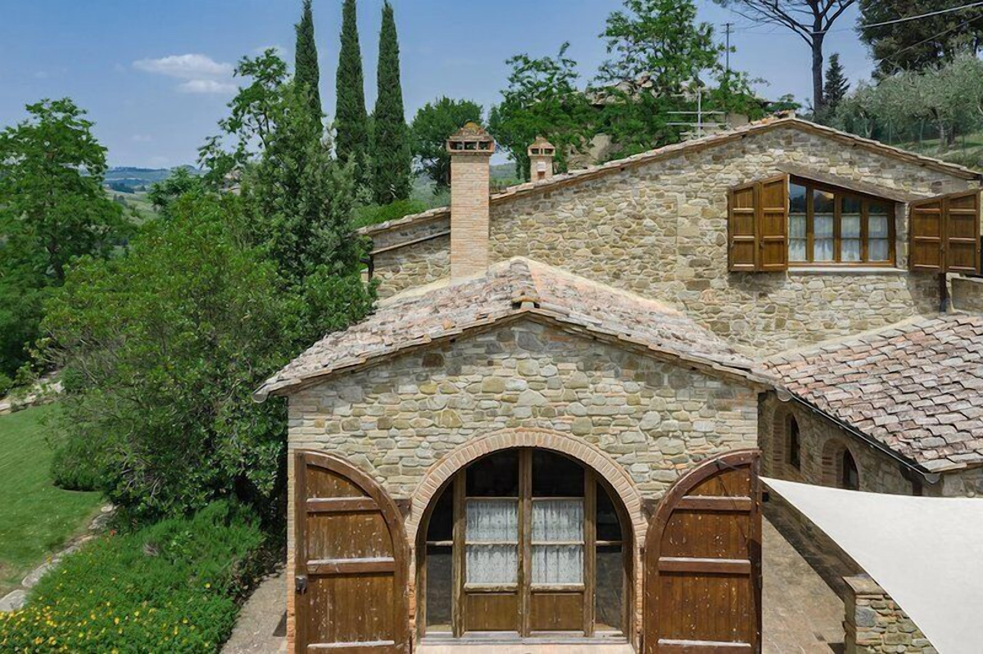 A stone house with arched windows and wooden shutters, surrounded by greenery and hills.