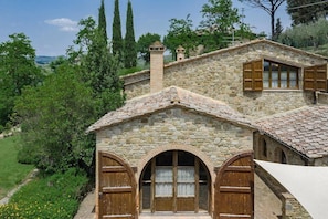 A stone house with arched windows and wooden shutters, surrounded by greenery and hills.