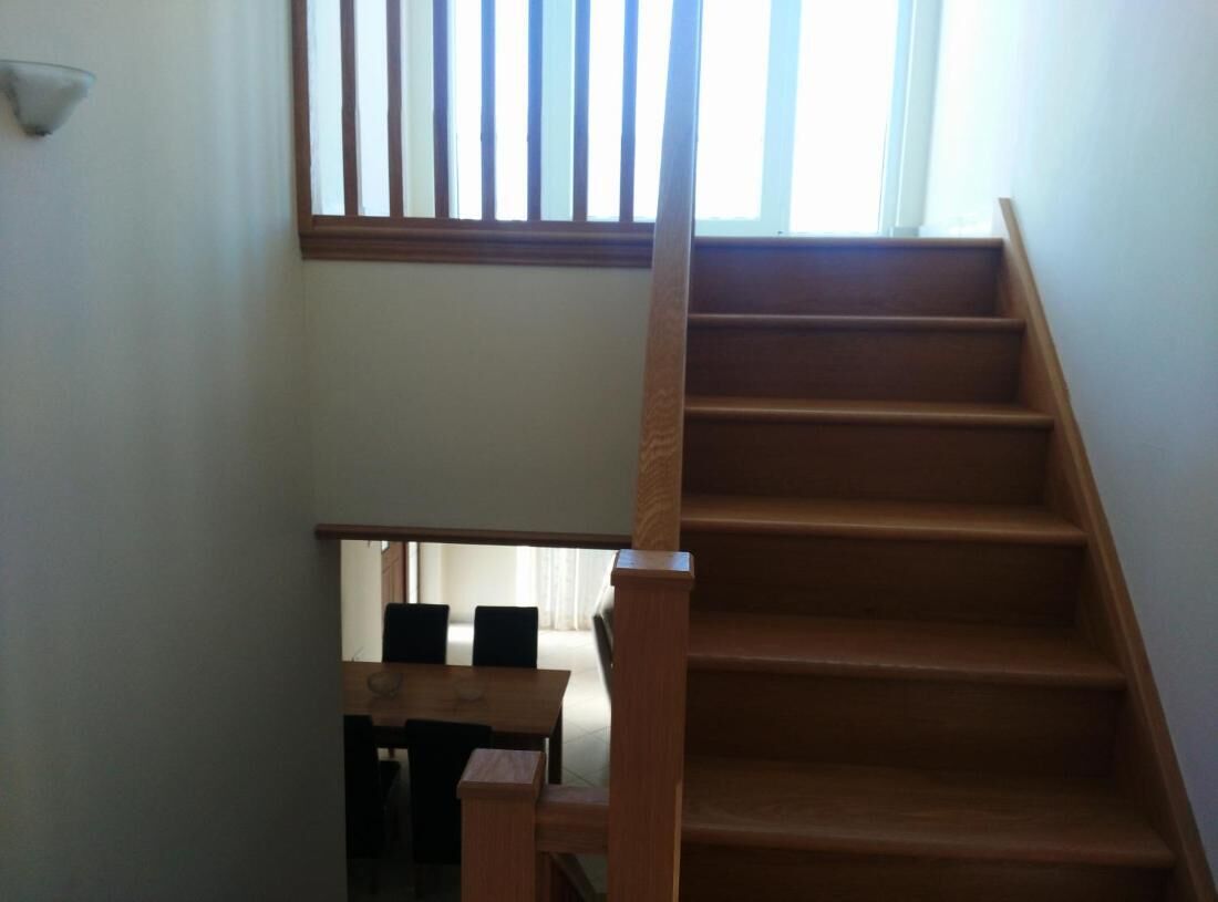 A wooden staircase with a handrail, leading to a lower level with a wooden table and chairs.
