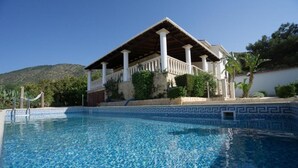 Ein Schwimmbecken mit gefliestem Rand, ein überdachter Außenbereich mit Säulen und eine Berglandschaft im Hintergrund.