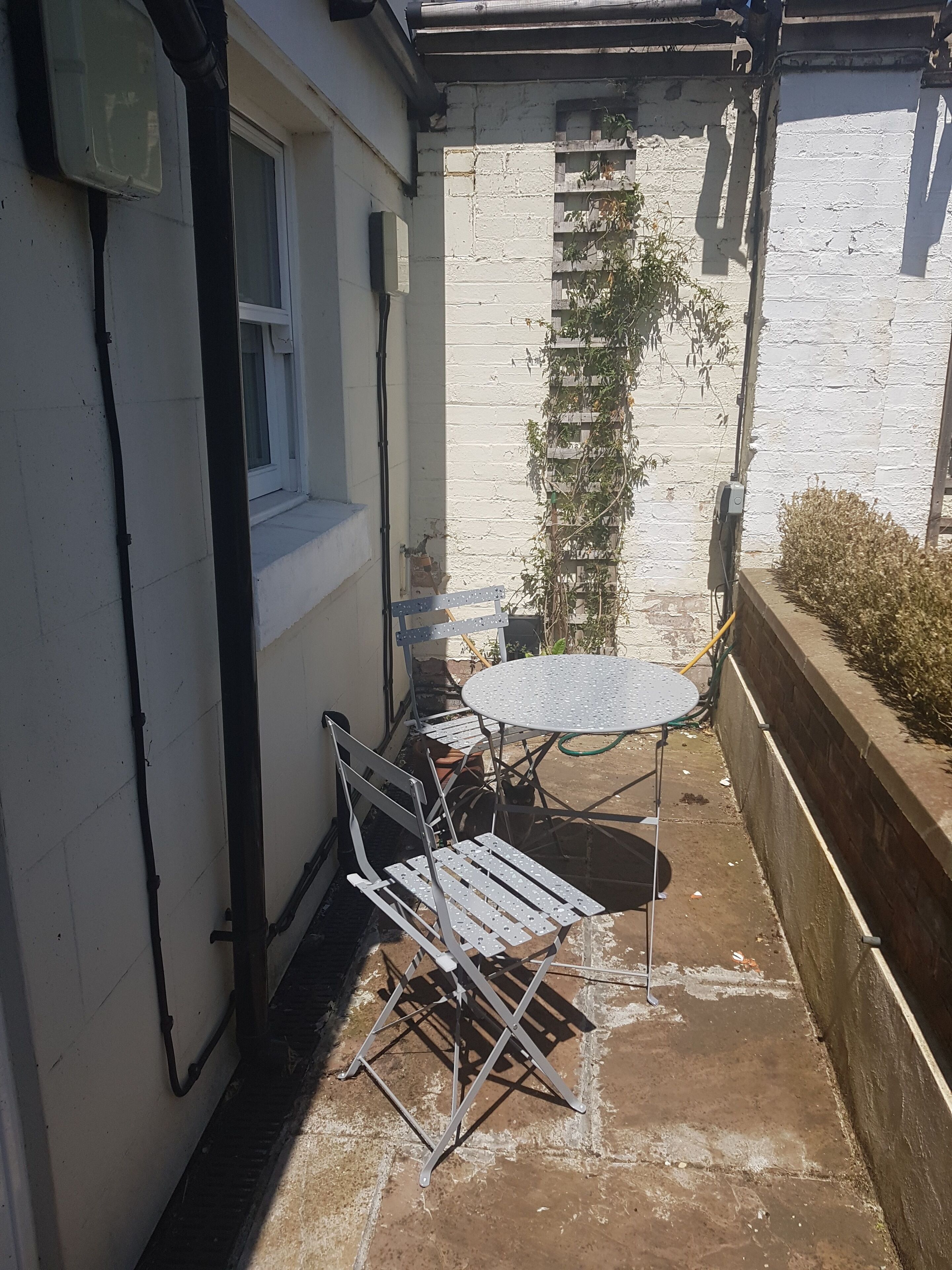 A small patio with a metal table and chairs, a window, and a brick wall with climbing plants.