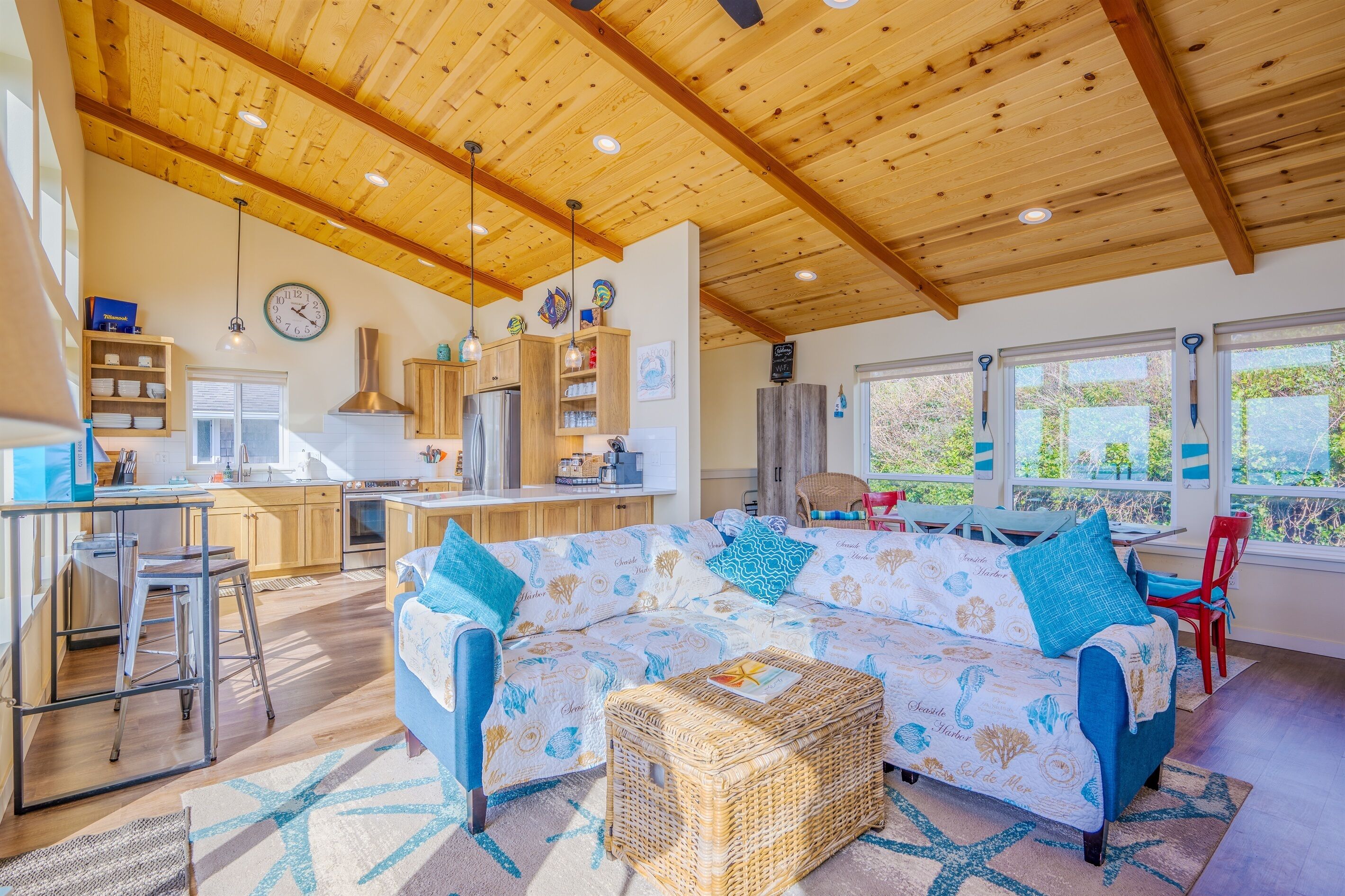 A spacious living area with a wooden ceiling, a sofa with patterned upholstery, a wicker ottoman, and a kitchen in the background.