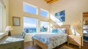 Un dormitorio con una cama grande, dos mesitas de noche, un escritorio y una silla. Tres ventanas ofrecen vistas al exterior.