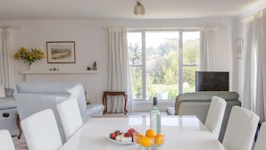 Zona pranzo con tavolo bianco, sedie e una ciotola di frutta. Sullo sfondo, un divano e una televisione.