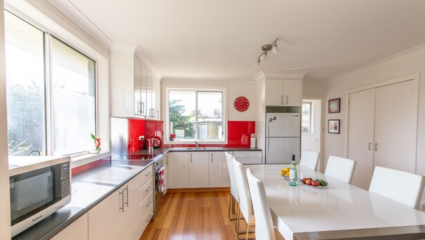 Una cucina moderna con mobili bianchi, uno schienale rosso, un forno a microonde integrato e un'area pranzo con sedie e tavolo bianchi.