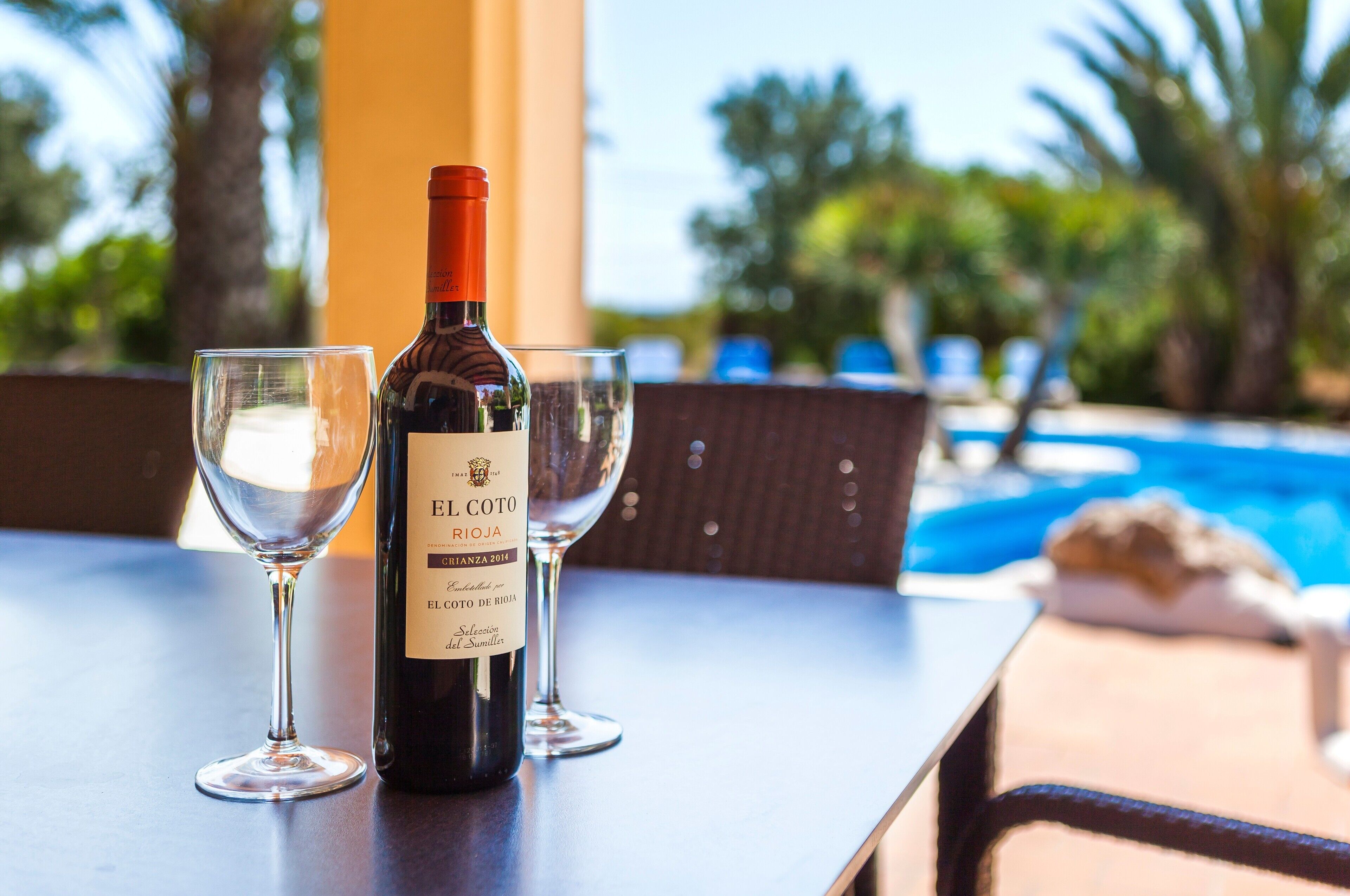 A bottle of El Coto de Rioja wine and two glasses on a table with a pool view.