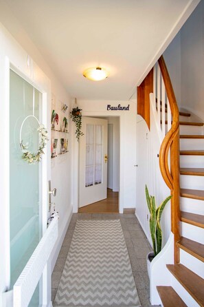 Un couloir avec un escalier en bois, un radiateur, un tapis à motifs en chevrons et une porte portant l’inscription « Bauland ».