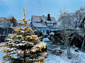 Une maison recouverte de neige, avec une cheminée bien visible, un arbre enneigé au premier plan et un ciel dégagé.