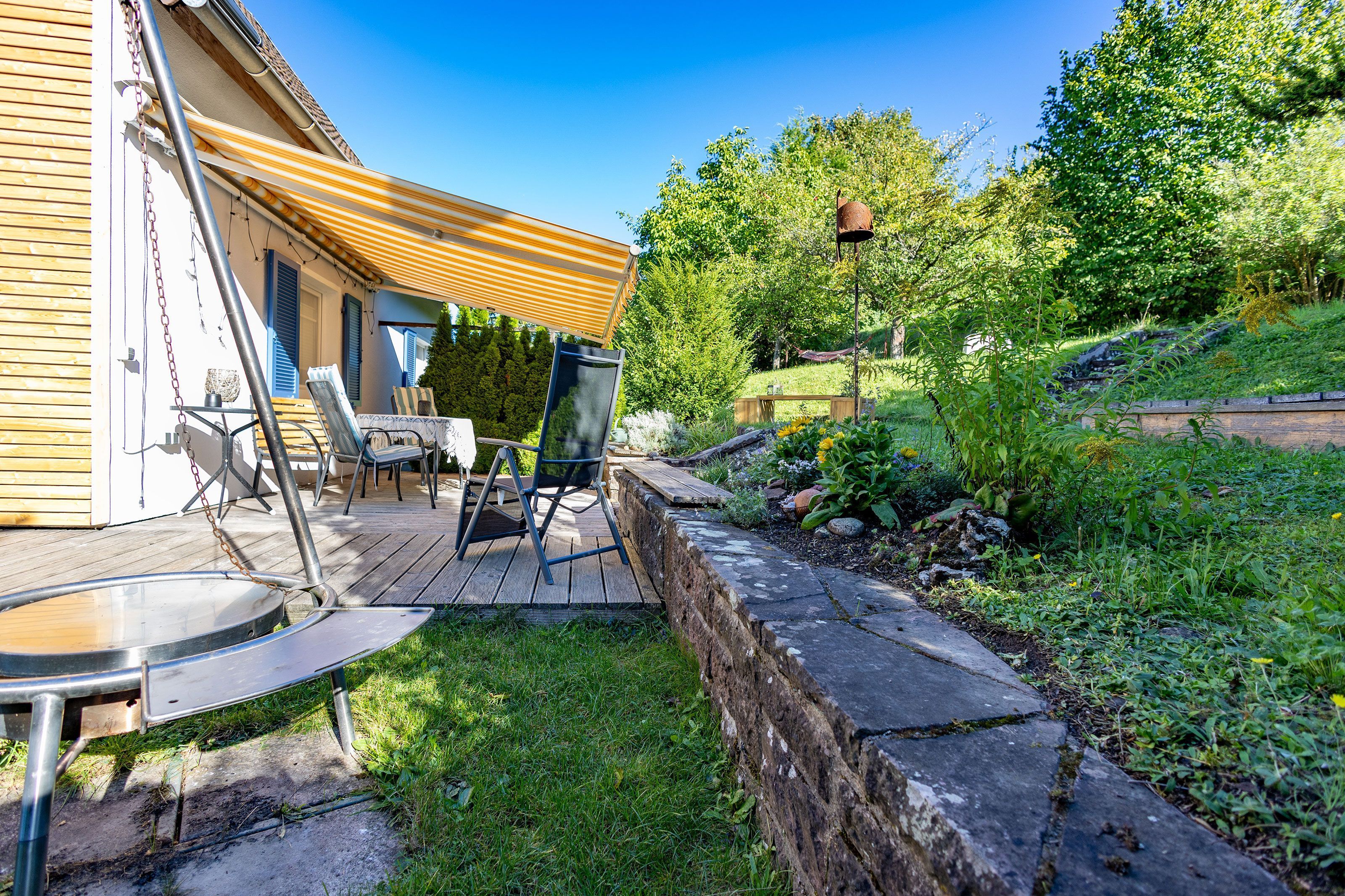 Une terrasse en bois avec des meubles d’extérieur, un mur de soutènement en pierre et un jardin agrémenté de diverses plantes et d’une clôture en bois.