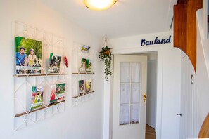 Un couloir avec une porte blanche portant l’inscription « Bauland », un mur blanc orné d’une étagère géométrique où sont posés des livres et des magazines, et une plante suspendue.