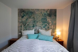 A neatly made bed with a metal headboard, a bedside lamp, and a floral-patterned wall behind it. - Bauland Vacation Apartment - Paula's Vacation Home   (Buchen)