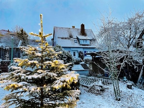 A snow-covered house with a visible chimney, a snow-covered tree in the foreground, and a clear sky.