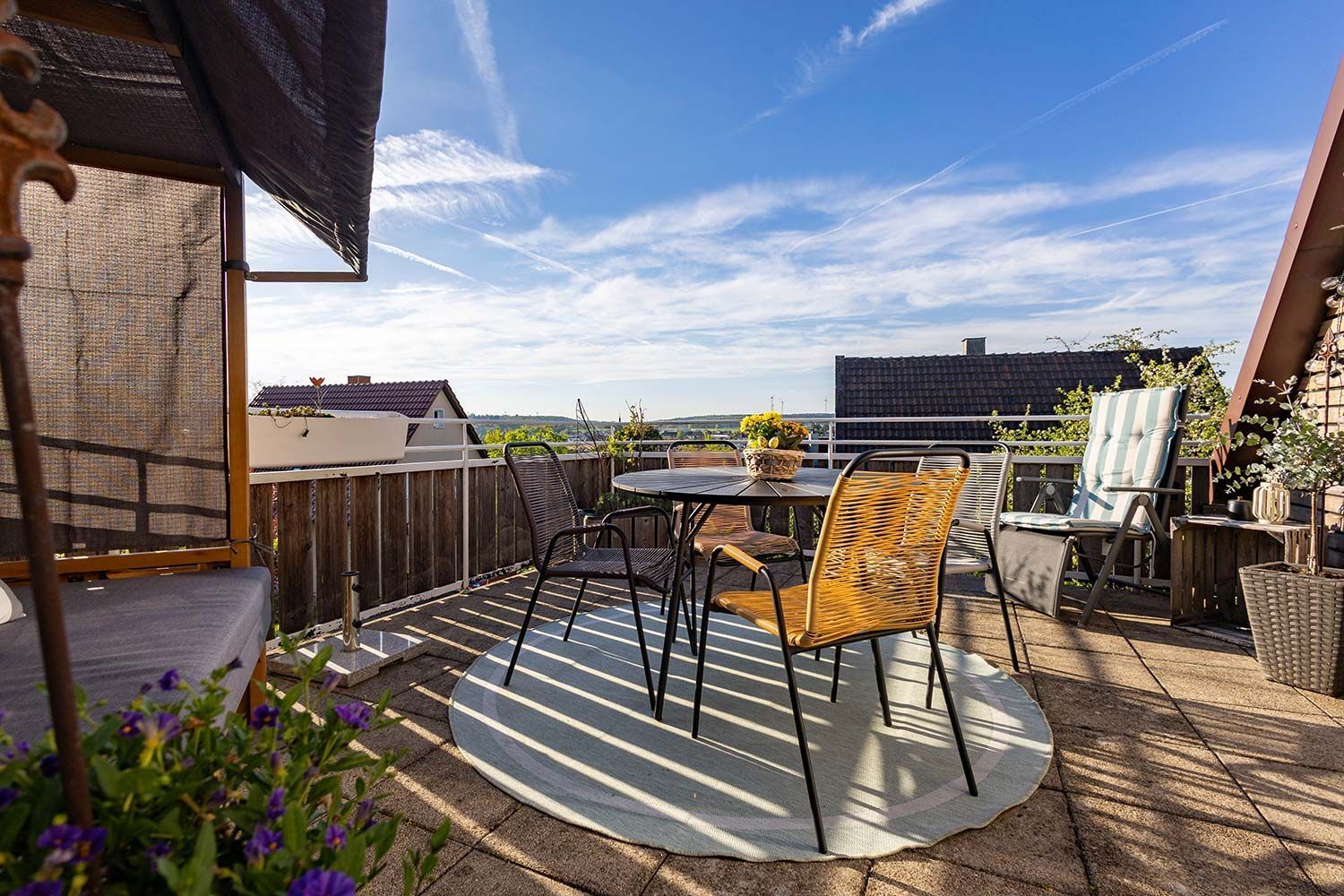 Une terrasse sur le toit, avec une table et des chaises, un fauteuil en osier, une petite table avec un panier, et une vue sur le paysage urbain.