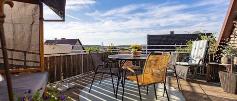 Une terrasse sur le toit, avec une table et des chaises, un fauteuil en osier, une petite table avec un panier, et une vue sur le paysage urbain.