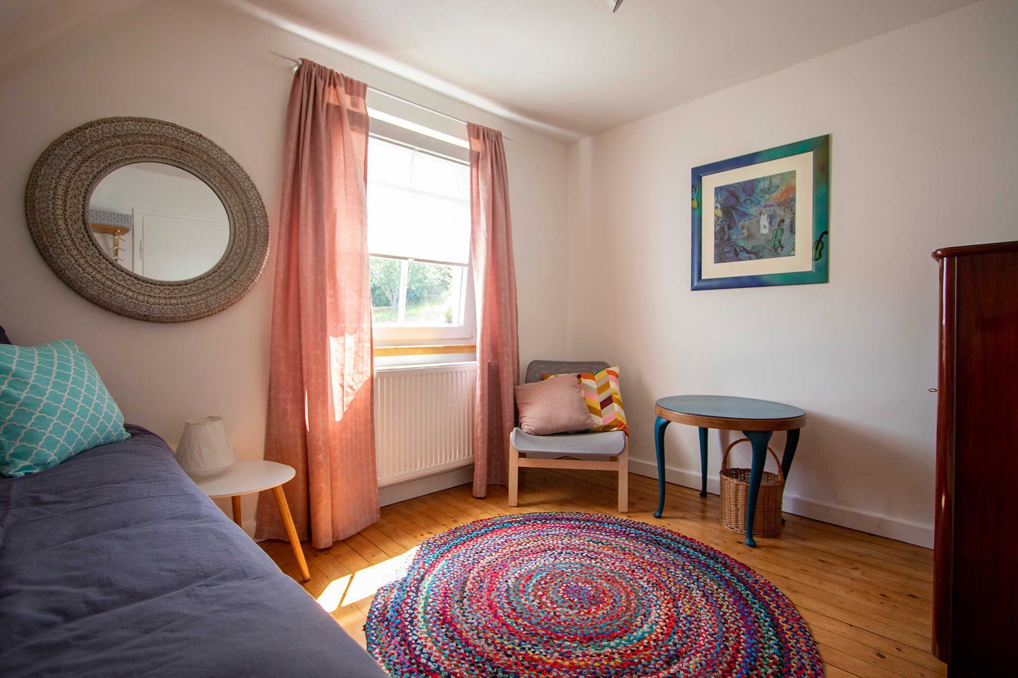 Ein Schlafzimmer mit Bett, Sessel, kleinem Tisch, rundem Spiegel, farbenfrohem Teppich und einem Gemälde an der Wand.