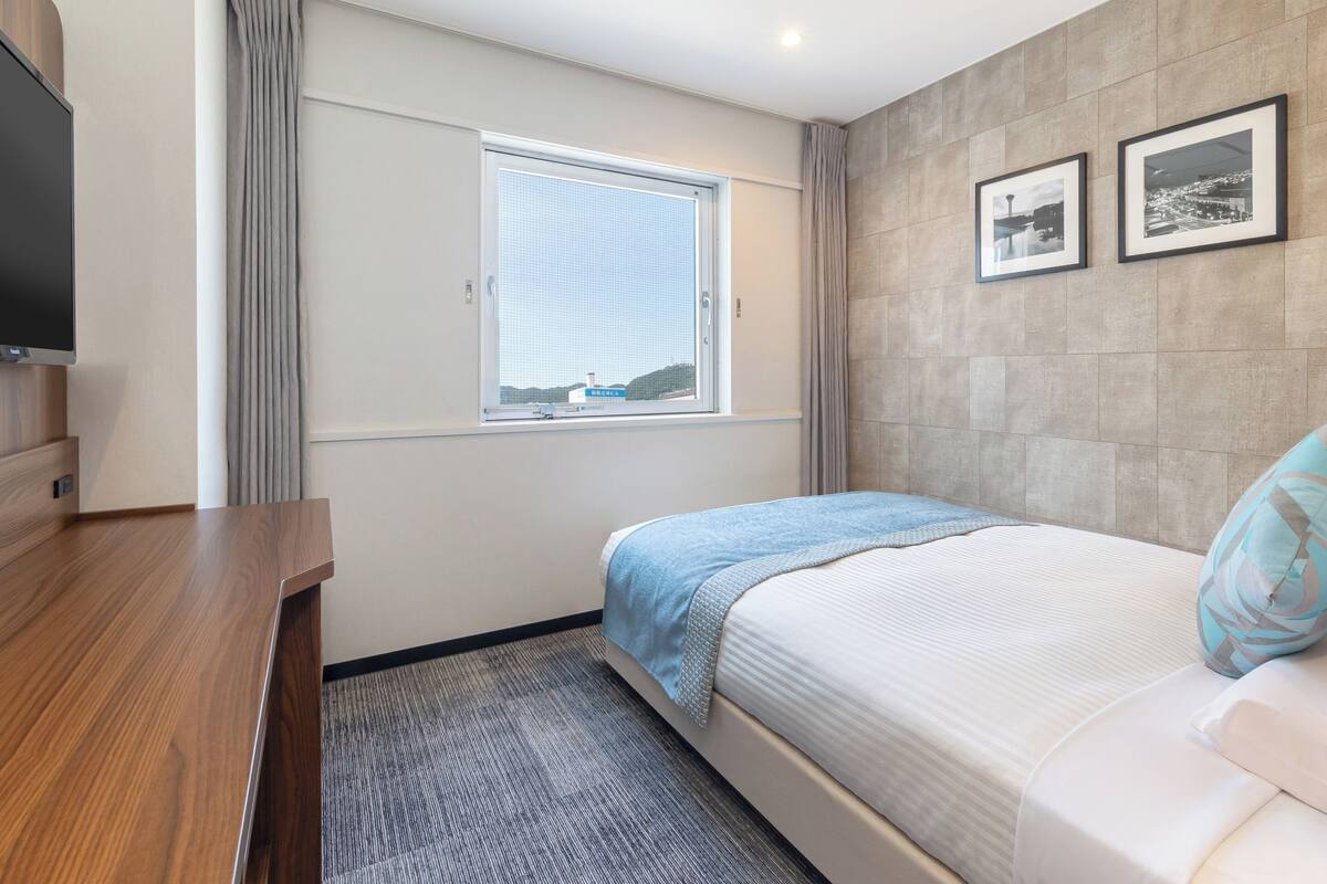 A modern hotel room with a bed, a desk, a TV, a window with curtains, and two framed pictures on the wall.