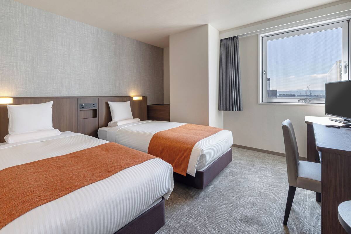 A hotel room with two beds, a desk, a chair, a TV, and a window with a view of the city.