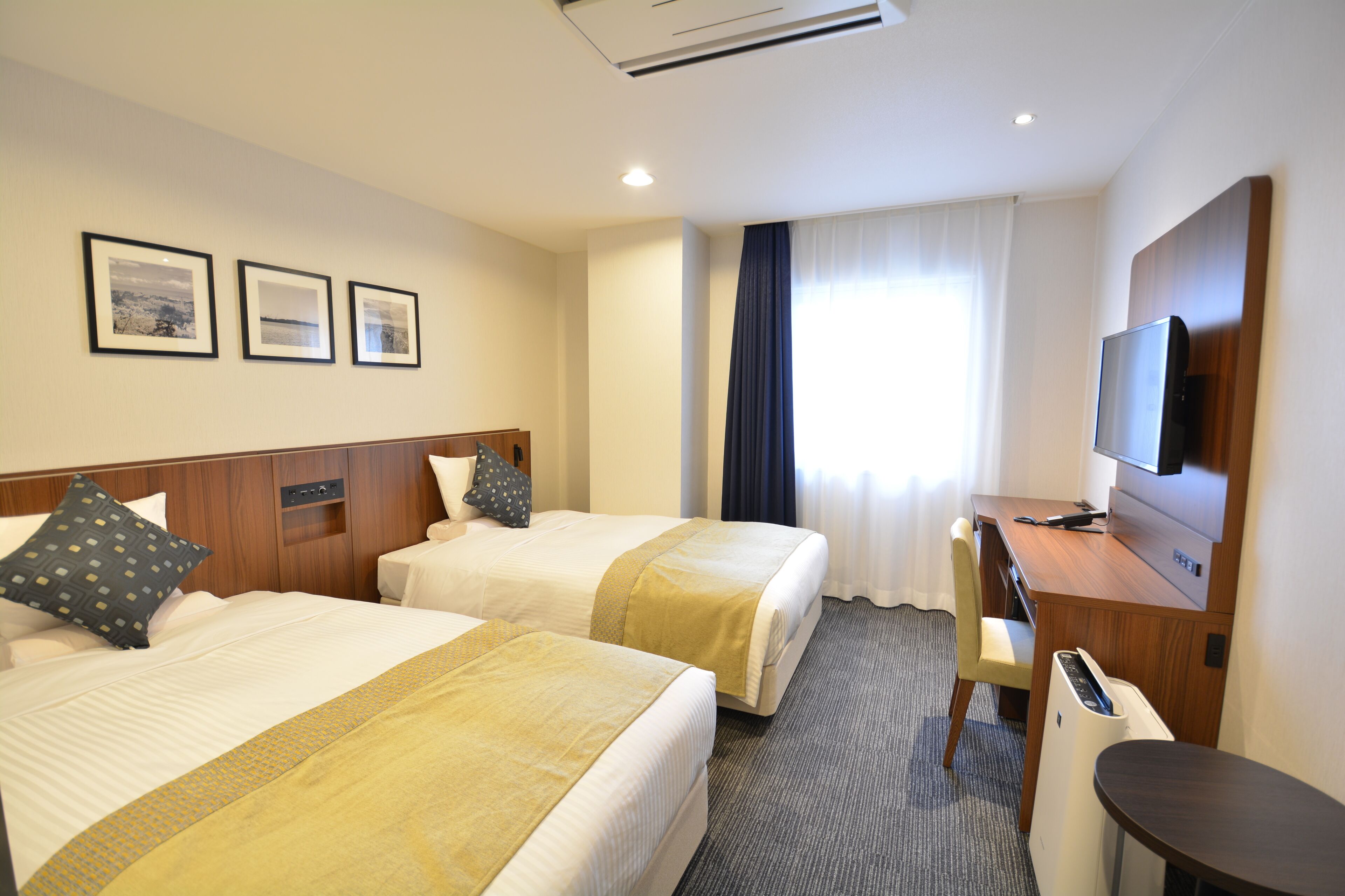 A hotel room with two beds, a desk, a chair, a TV, and a window with curtains.