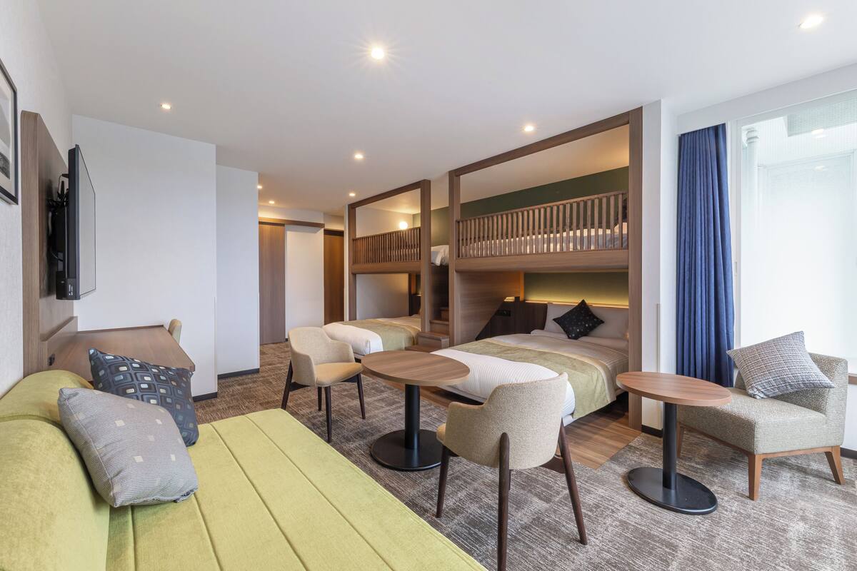 A modern hotel room with a sofa, armchairs, a round table, a bunk bed, and a TV.