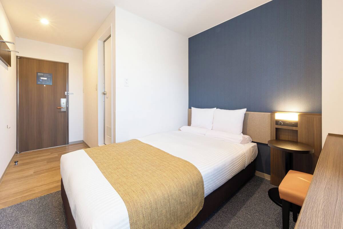 A hotel room with a bed, a desk, a chair, and a nightstand with a lamp.