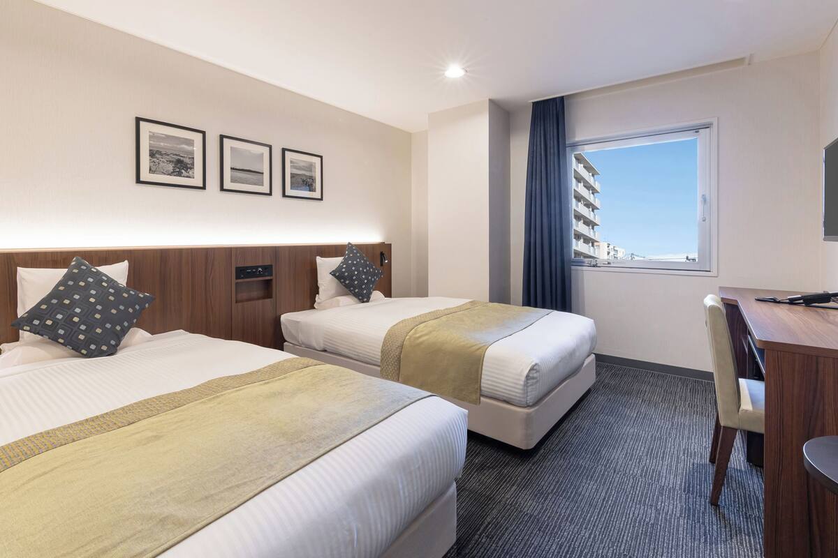 A hotel room with two beds, a desk, and a view of a building through a window.