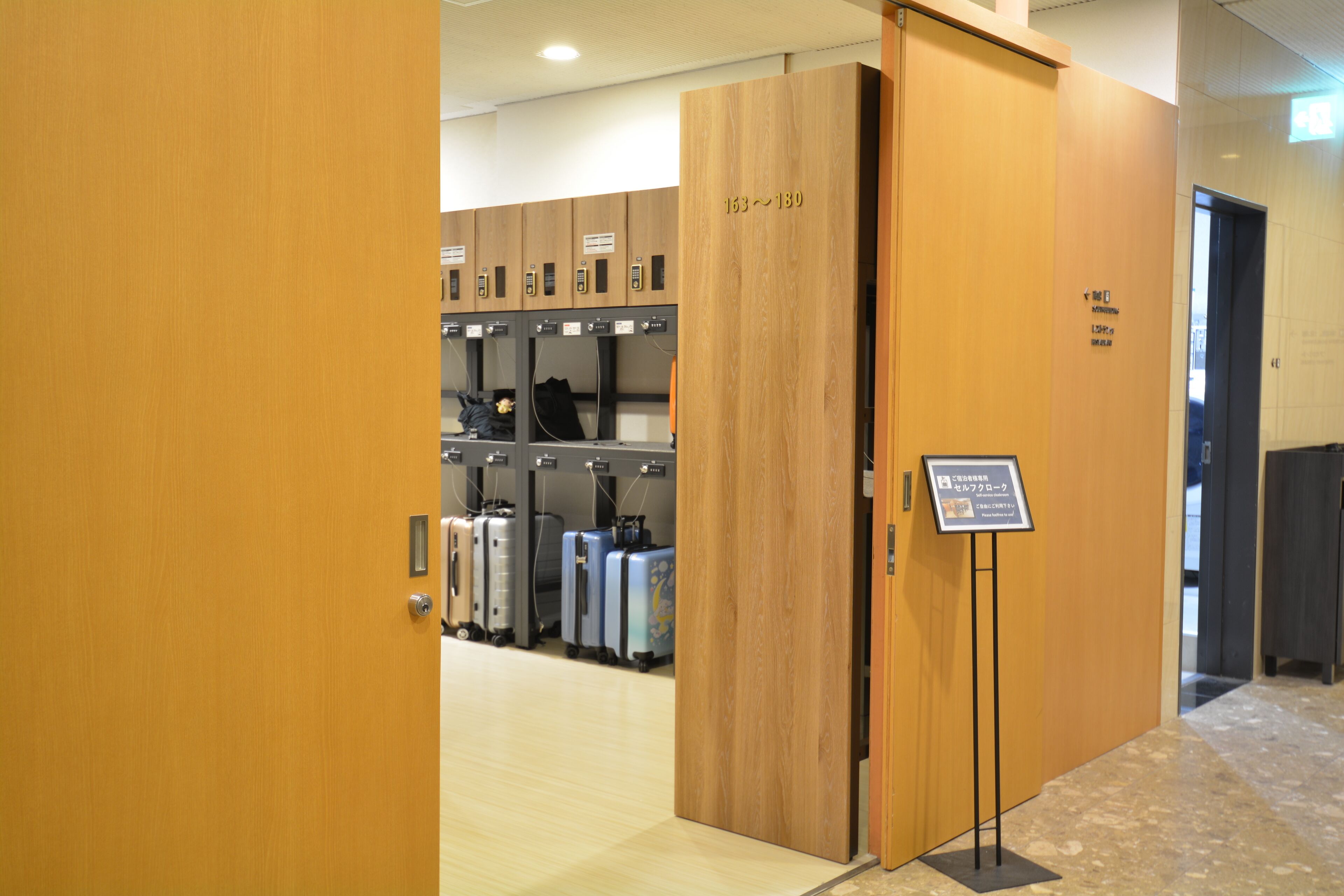 A hotel room with a wooden door, a sign indicating room number 163-180, and a row of suitcases in the background.