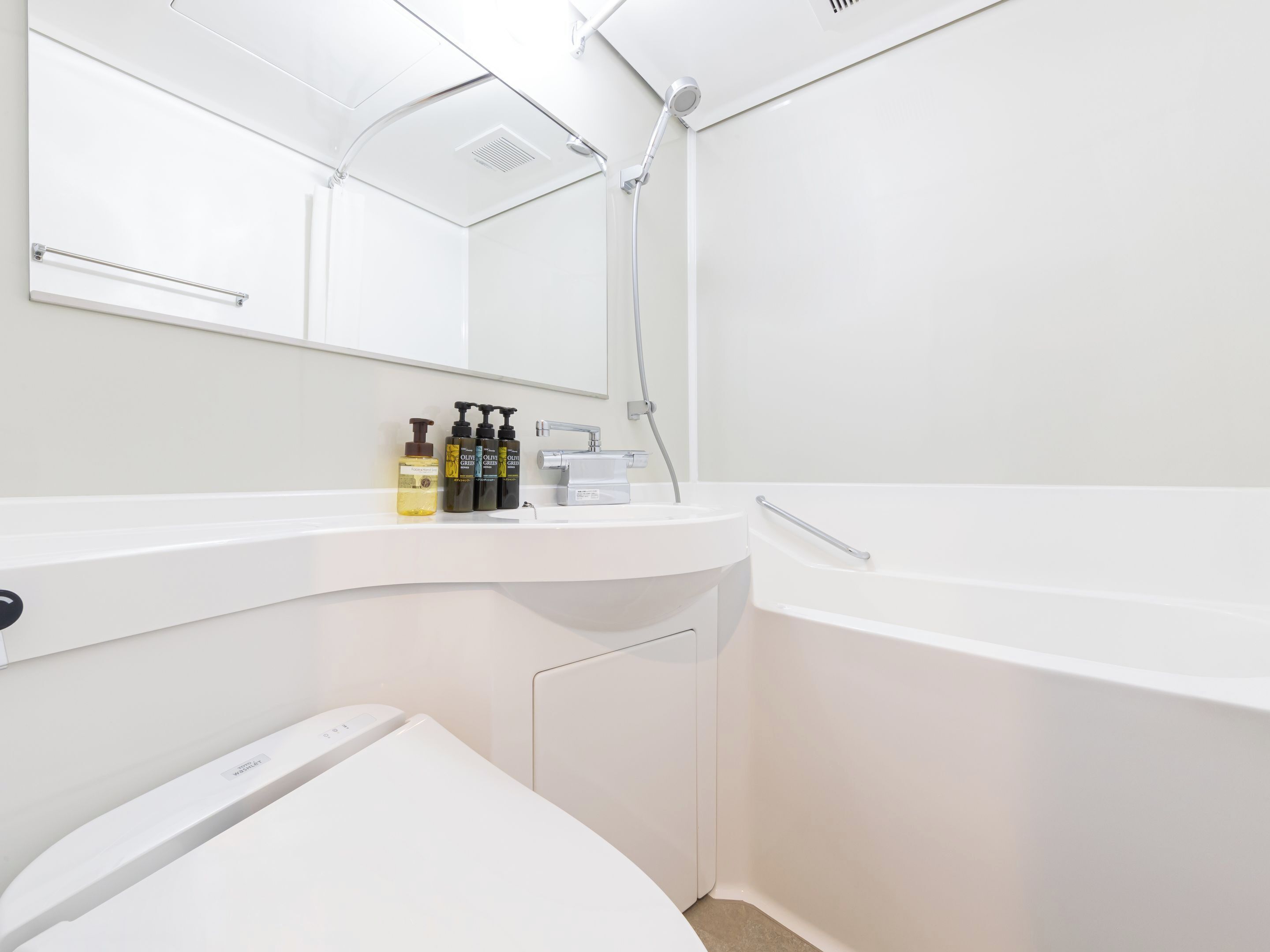 A modern bathroom with a white sink, a mirror, and a shower. There are three bottles of olive oil on the sink.