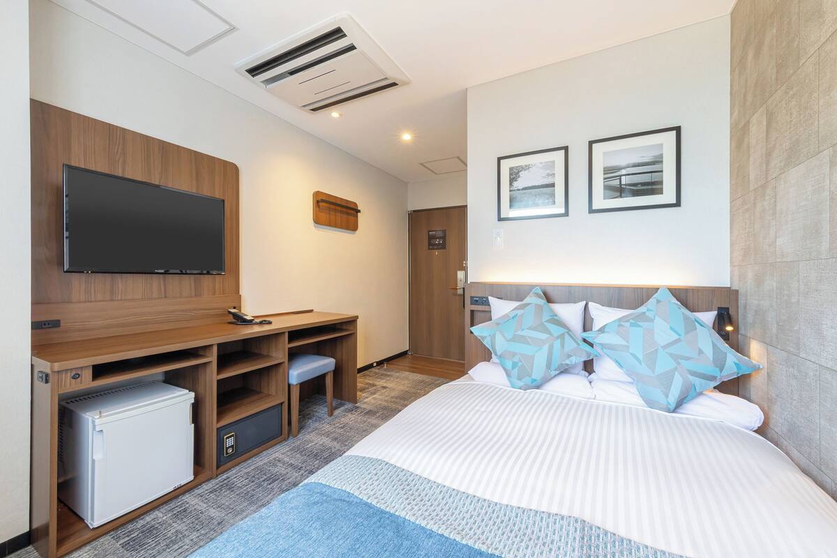 A modern hotel room with a flat-screen TV, a wooden desk with a mini-fridge, a bed with patterned pillows, and a wall-mounted air conditioner.