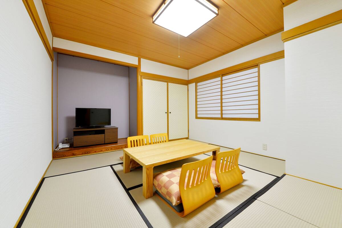 A traditional Japanese room with tatami flooring, a wooden table, yellow cushions, a TV on a low wooden stand, and shoji screens.