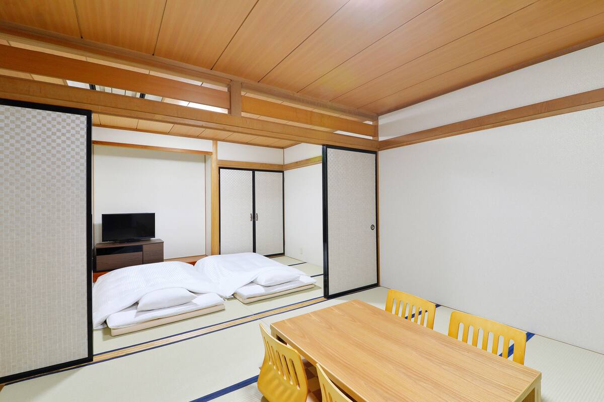 A traditional Japanese room with tatami flooring, a wooden table, yellow chairs, and a television on a low shelf.