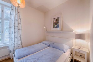 Une chambre à coucher comprenant un lit, une table de chevet, une lampe, une fenêtre avec des rideaux et une étagère murale ornée d’un tableau.