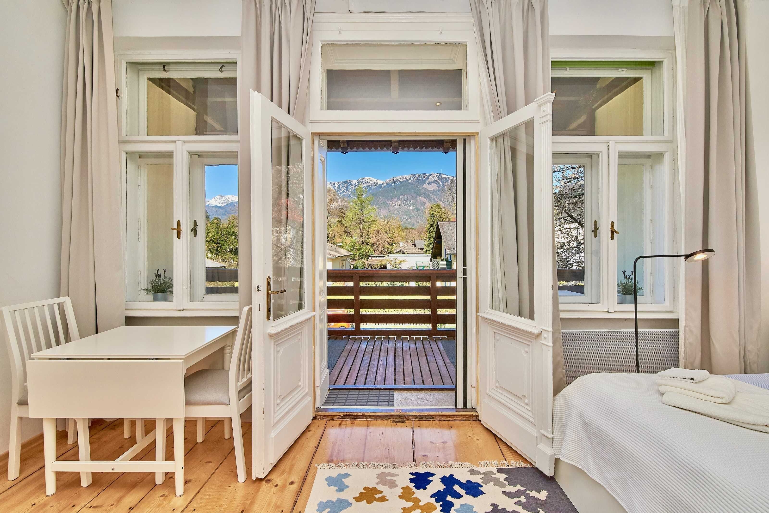 A room with a bed, a desk, and chairs, a window with a view of mountains, and a balcony with a wooden deck.
